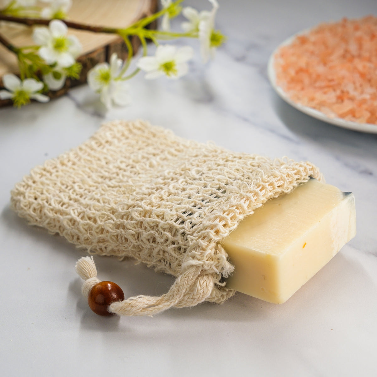 Natural soap bar with a loofah sponge on a light surface with flowers and salt in the background.