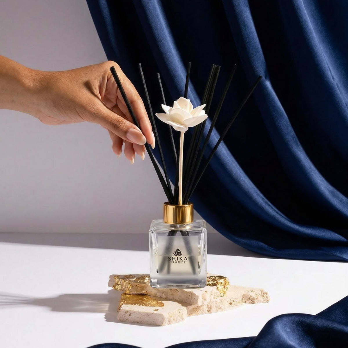 Hand holding a diffuser with a white flower on top against a blue curtain background