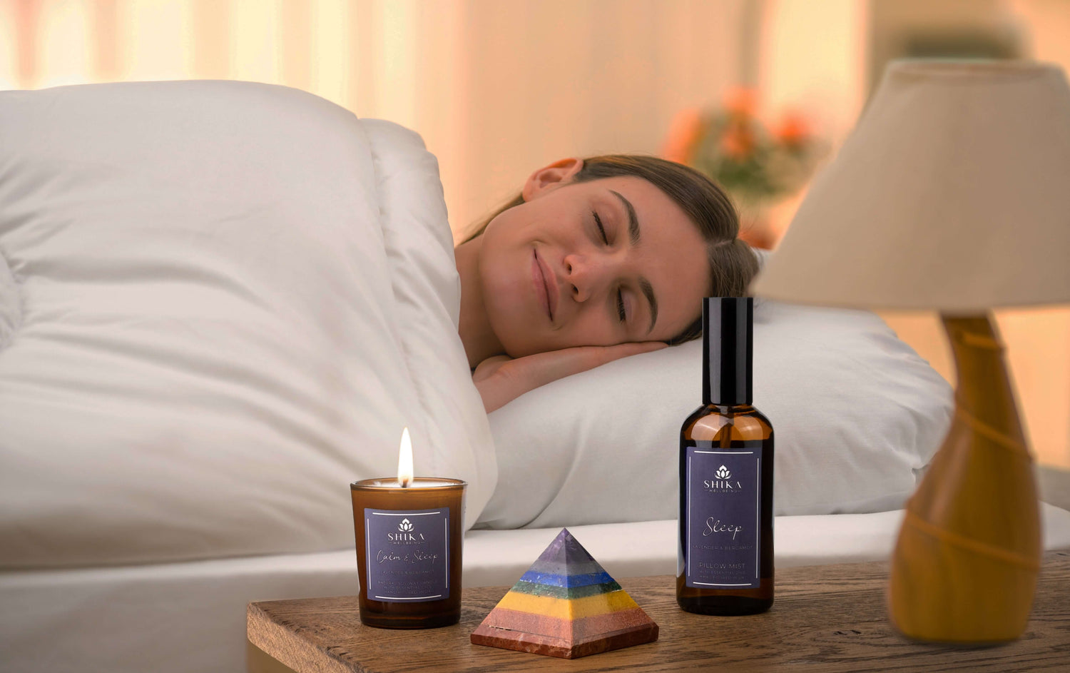 Woman lying in bed with her eye closed. There is a bedside table with the Shika Wellbeing Sleep gift set products on there and a lamp. The products are a bottle of sleep spray, a sleep candle and a pyramid crystal