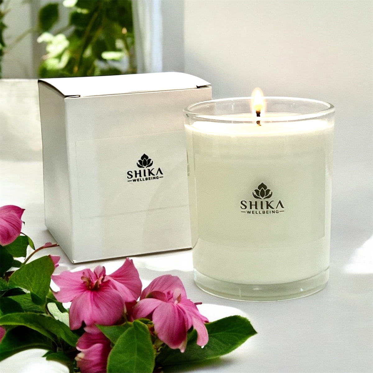 Shika Wellbeing candle with a box and pink flowers on a white surface